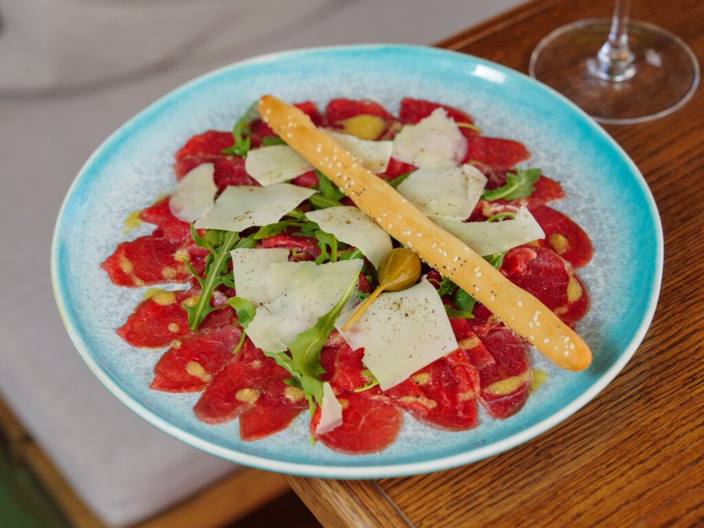 Close-up of carpaccio served with arugula, cheese, and a breadstick on a blue plate.
