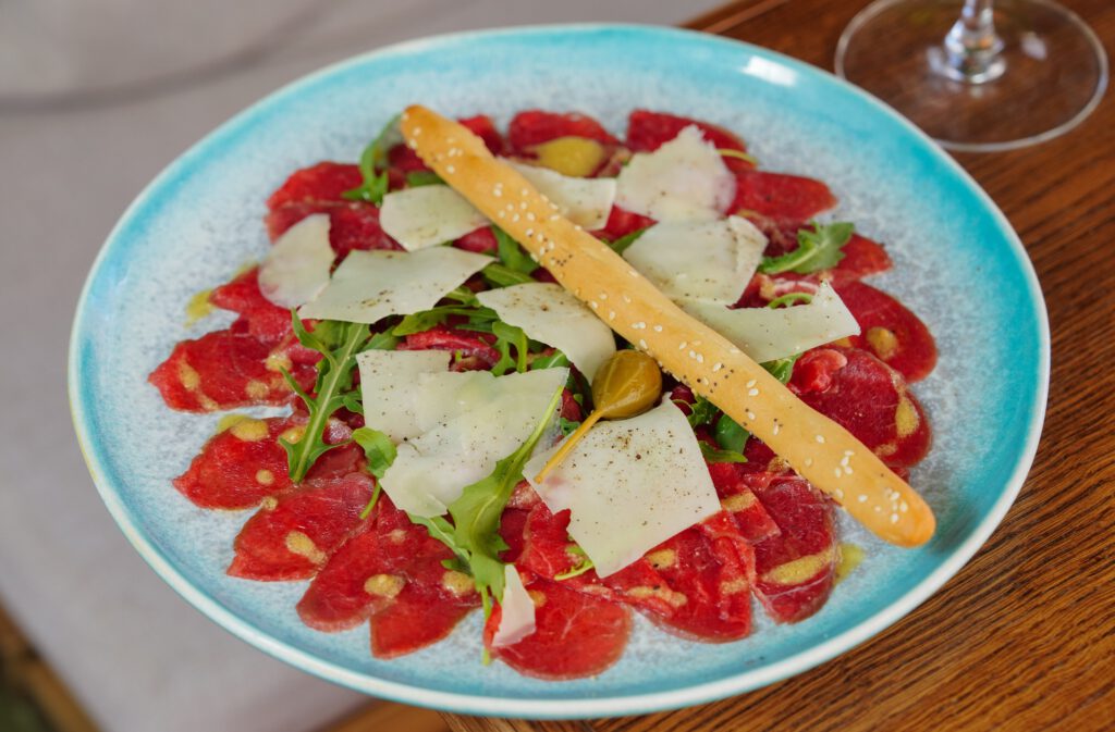 Close-up of carpaccio served with arugula, cheese, and a breadstick on a blue plate.